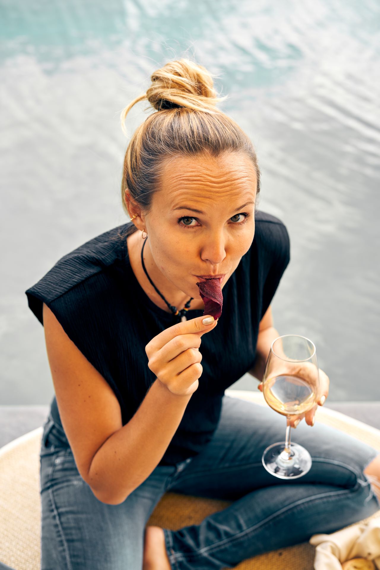 Junge Frau mit Dutt in Jeans sitzt am Wasser mit einem Weißwein in der Hand und isst lila Chips während sie in die Kamera schaut.