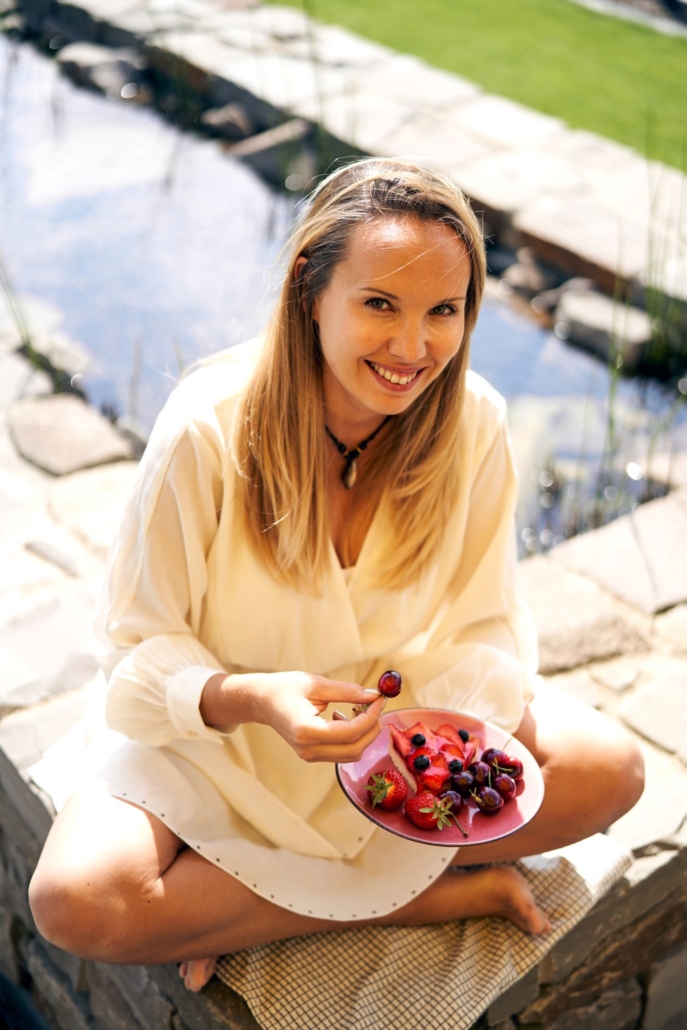 Schlanke blonde Frau isst Beeren und Kuchen im Schneidersitz in einem beigen Kleid auf einer Teichmauer und lächelt.