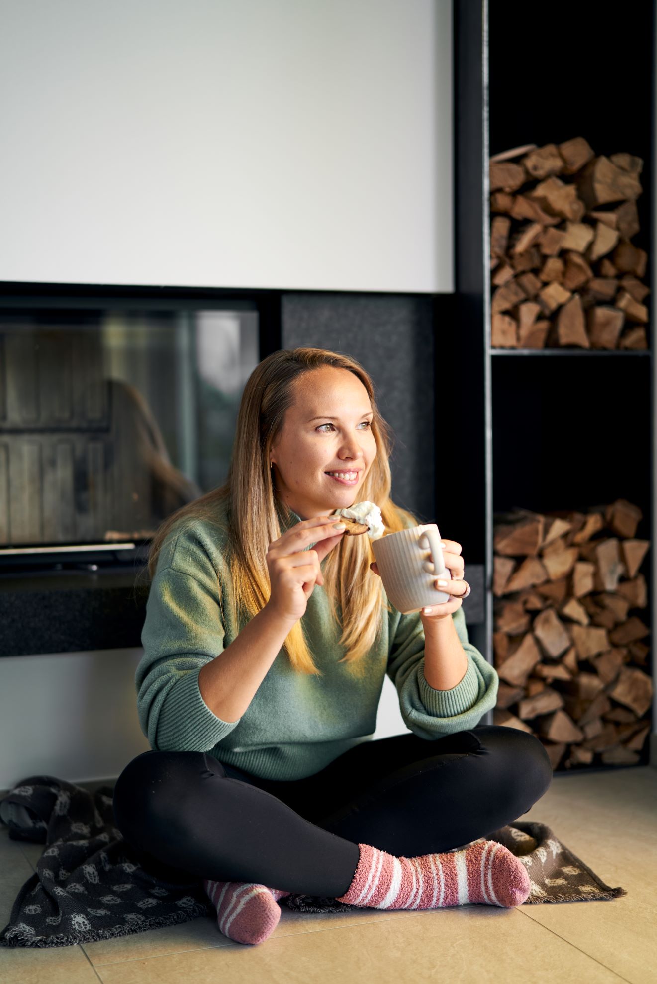Schlanke Frau im Schneidersitz vor dem Kamin genießt eine heiße Schokolade