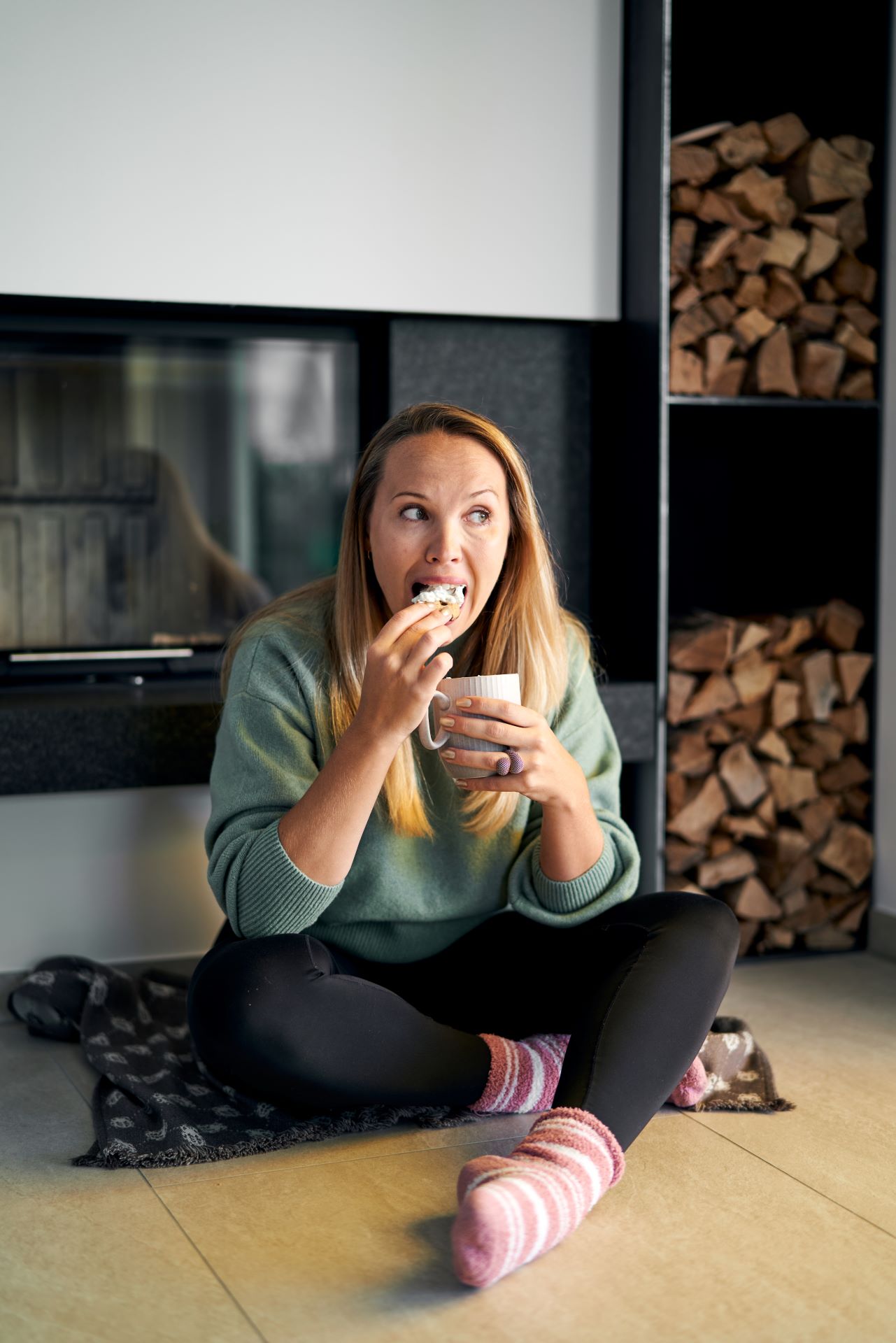 Frau im Schneidersitz vor einem Kamin beißt mit voller Freude in einen Kecks mit Sahne und freut sich diesen zu essen.