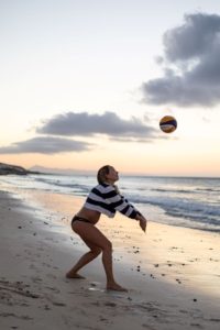 Schwangere Frau im Bikini und gestreiften lockeren Pulli spielt Beachvolleyball am Strand